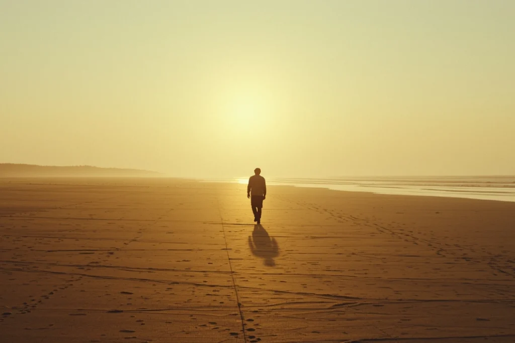 Back-facing person walking forward across wide open beach at dawn, upright steady posture, soft golden morning light and expansive sky ahead