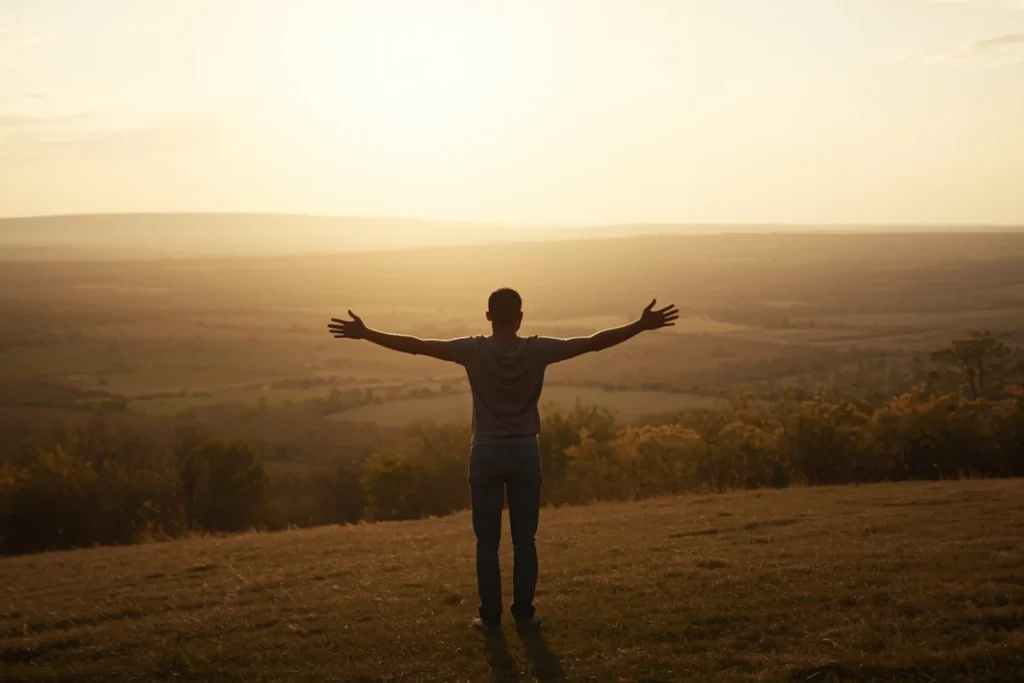 Person standing at hilltop crest at dawn, looking over broad landscape, warm morning light, sense of emergence