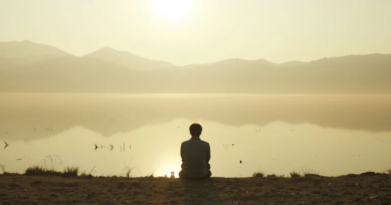 Person seated at edge of still lake at dawn, back-facing, calm water reflecting soft golden morning light, wide open landscape
