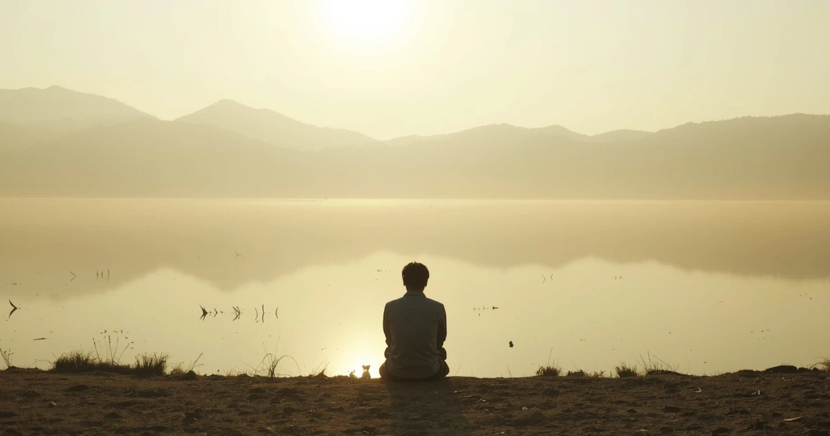 Person seated at edge of still lake at dawn, back-facing, calm water reflecting soft golden morning light, wide open landscape