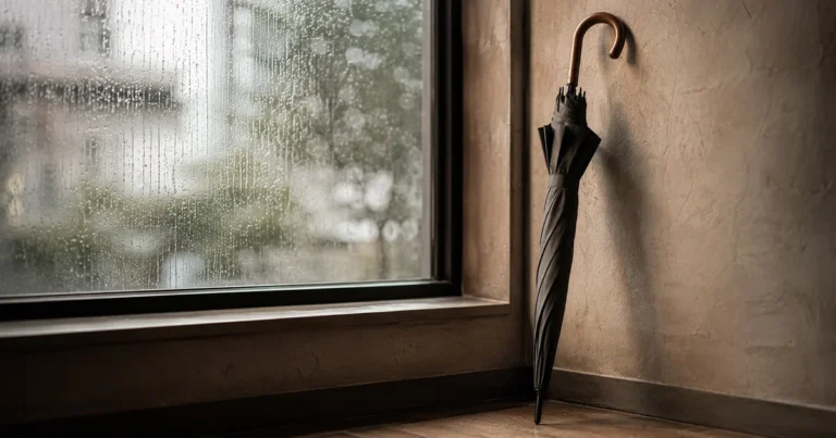 A single unopened umbrella leaning against a wall near a rain-streaked window in soft gray daylight