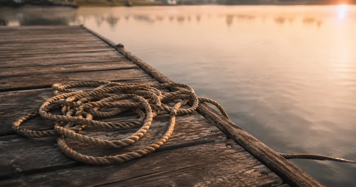 A length of rope coiled loosely on a wooden dock with one end trailing into calm water in soft morning light