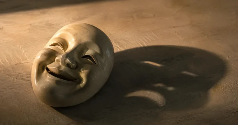 A smiling ceramic mask resting face-up on a table, its shadow cast in a different, tense expression, in soft evening light
