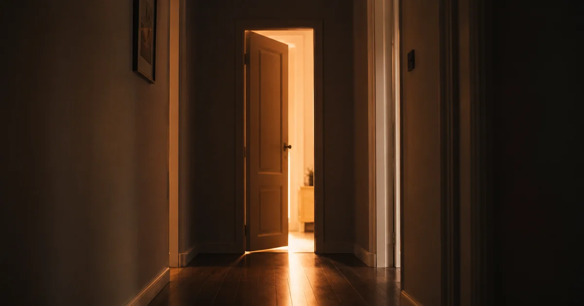 Empty hallway with a door left ajar, warm light spilling through into shadow