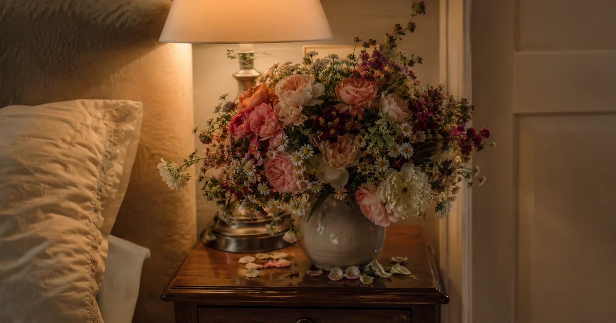 Overflowing bouquet crowding a small vase, petals spilling onto the nightstand, warm lamp light