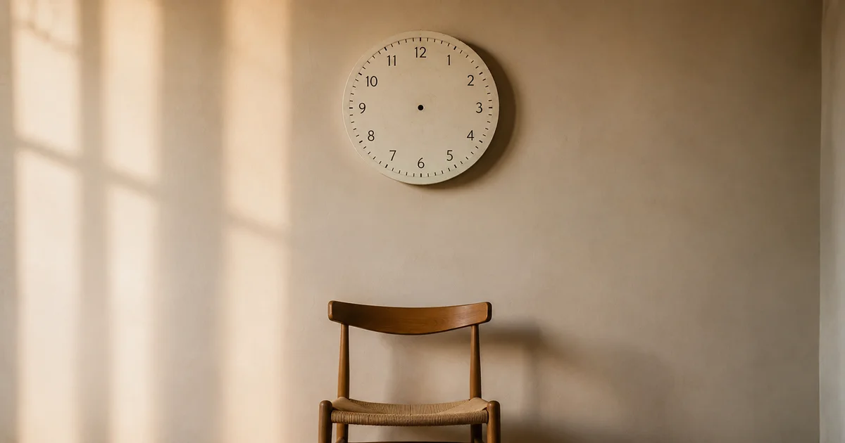 Clock with no hands on a plain wall, morning light crossing it, empty chair beneath