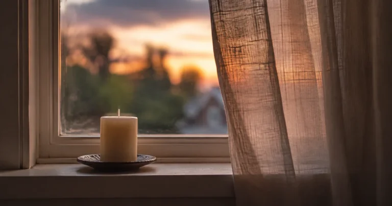 A single unlit candle on a windowsill with curtains slightly drawn, soft dusk light outside