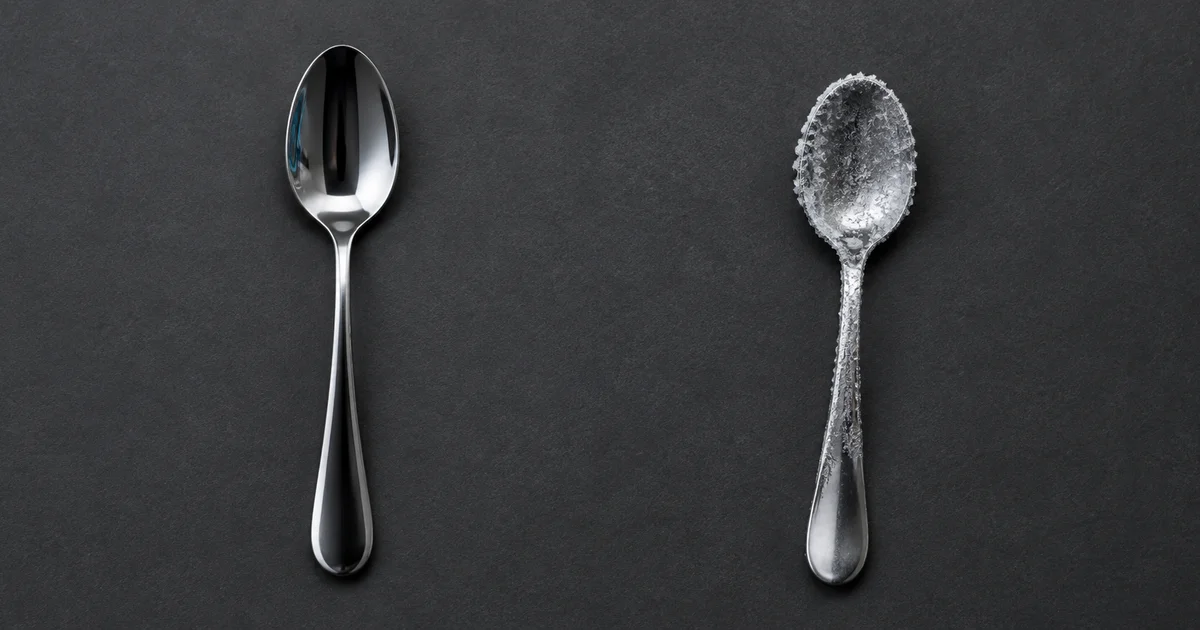 Two plain steel teaspoons on a dark gray surface, the left one clean and polished, the right one partly covered in frost