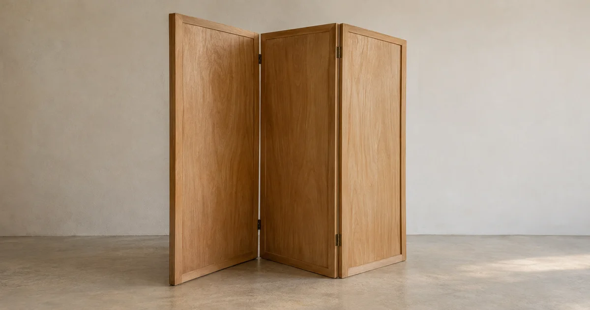 A plain three-panel wooden folding screen standing partly folded at an angle on a bare floor by a neutral wall.