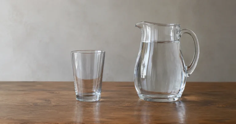 An empty glass standing beside a full pitcher of water on a plain table