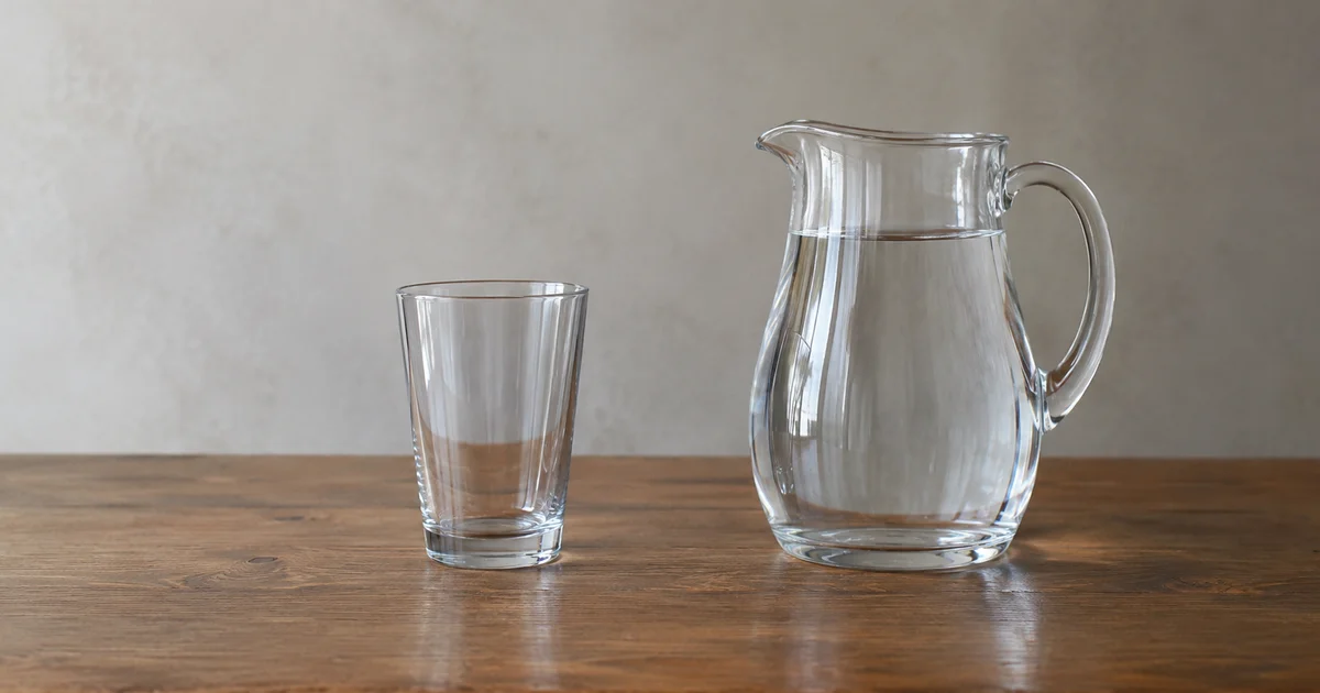 An empty glass standing beside a full pitcher of water on a plain table