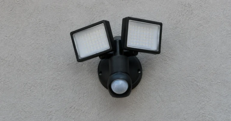 A security floodlight fixture mounted on a plain exterior wall, switched off, its sensor lens visible